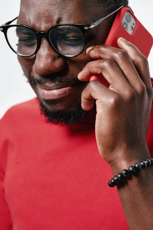 African man with dark skin wearing glasses and red shirt talking on smartphone with emotional expression closeup. Communication, technology and mobile phone use for connection and conversation.