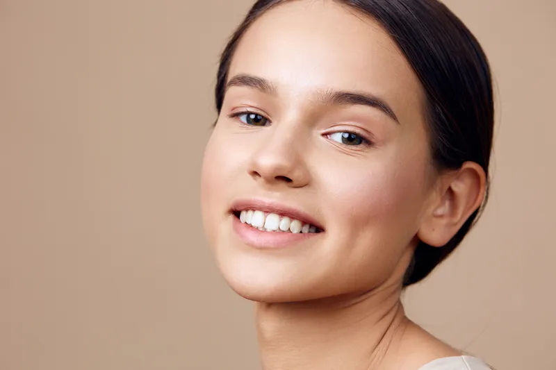 Young woman with a bright smile, showcasing natural beauty and healthy skin tone against a warm beige background, representing joy and positivity