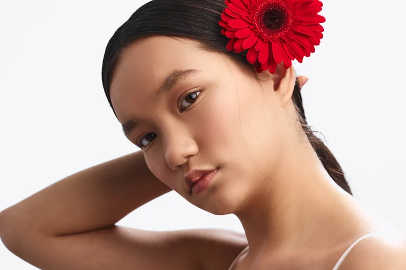 asian woman with a vibrant red flower in her hair gazes softly at the camera, revealing natural beauty and calm elegance in a simple studio portrait.