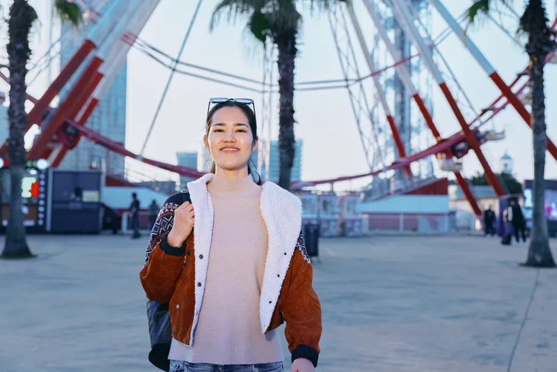 Woman walking outdoors in an urban plaza wearing a plush jacket over a soft sweater, smile on her face, sunglasses on head, modern arches and palm trees in the backdrop.