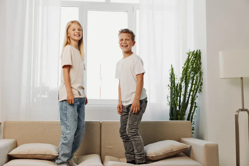 Happy children standing on a beige sofa in bright living room with large windows and indoor plant. Casual clothing and natural light create a relaxed, joyful home atmosphere for kids.