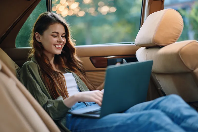 Young woman working on a laptop in a car, feeling relaxed and happy, wearing casual clothing, surrounded by a serene outdoor background with soft lighting.