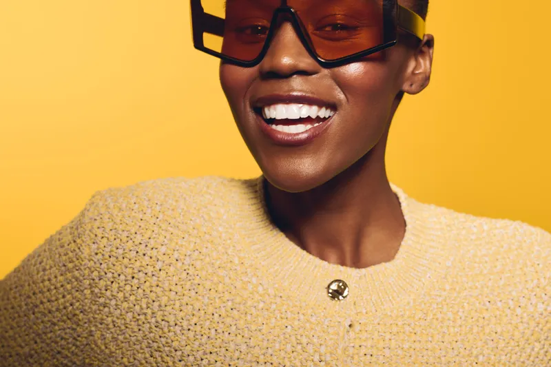 Smiling African American woman with dark skin wearing oversized sunglasses and a yellow knitted sweater on yellow background. Concept of fashion, beauty, and joyful mood.