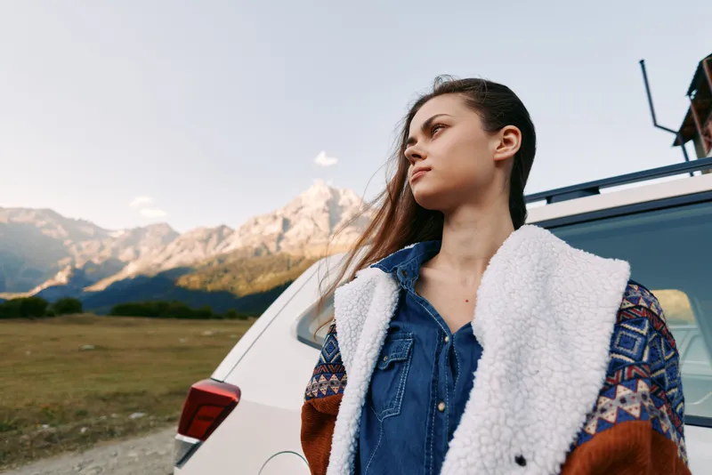 Woman portrait by car and mountains outdoors for travel and roadtrip, wearing jacket and denim coat. Young female gazes to horizon in scenic countryside with autumn sky and peaks.