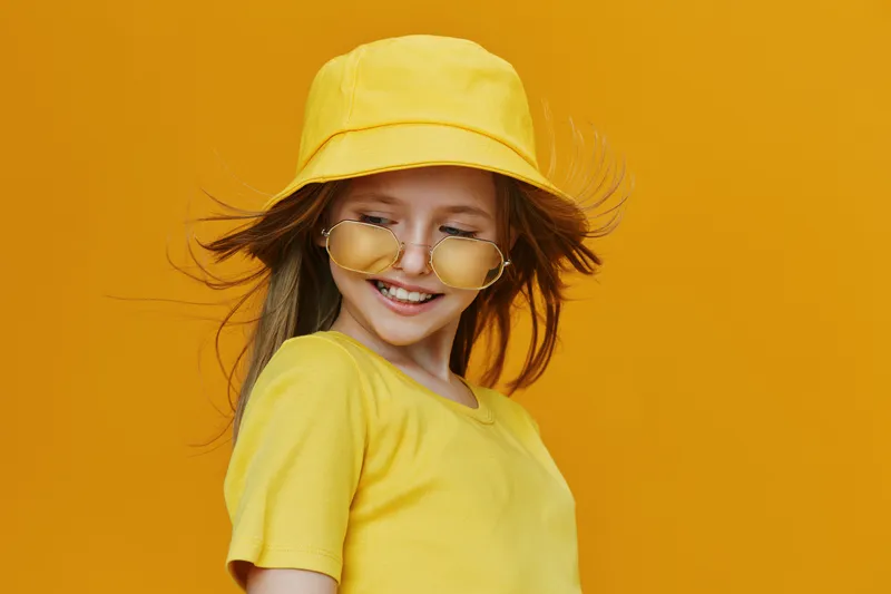 Young girl with long hair smiling in yellow summer outfit and hat against a vibrant yellow background, conveying joy and playfulness for seasonal lifestyle imagery