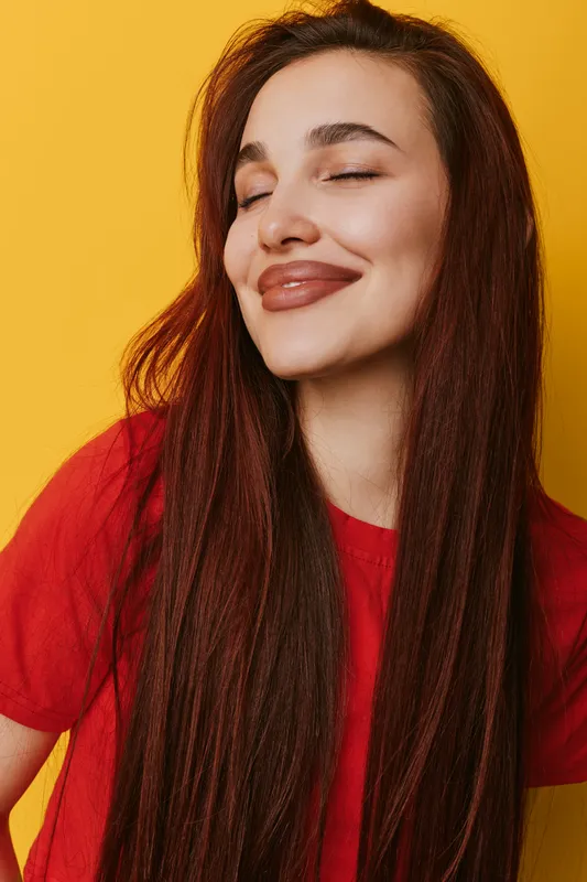 Young woman with long brown hair smiling with closed eyes wearing red shirt on yellow background. Portrait of happy female with natural makeup and relaxed expression in studio lighting.