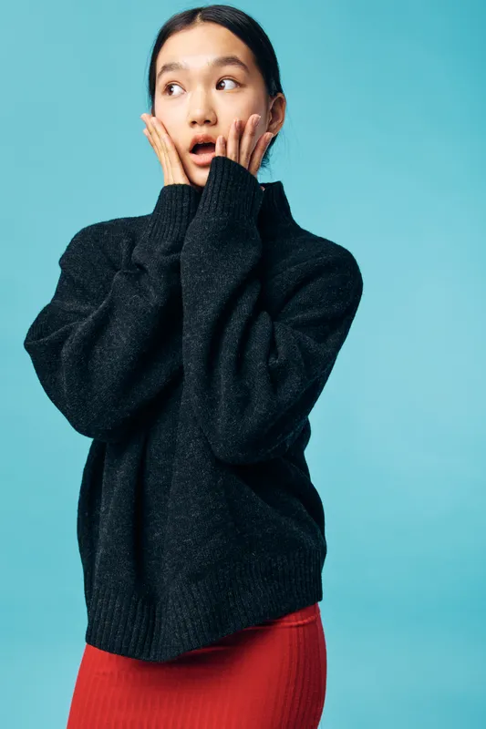 Woman with surprised expression holding face in hands wearing black sweater and red skirt looking away. Isolated on blue background with emotional reaction and casual outfit.
