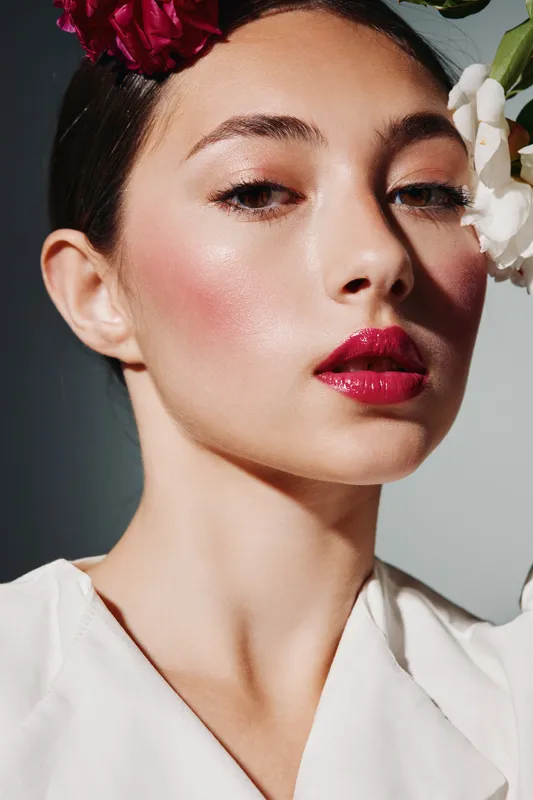 Close-up portrait of a stylish woman with glossy red lipstick and flawless glowing skin. Flower accessories along hairline highlight a creative beauty fashion concept in studio lighting.