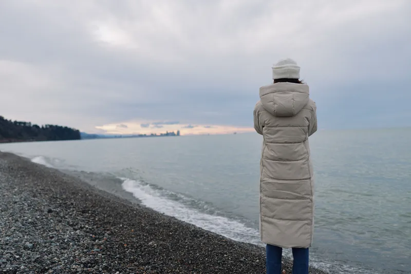 Coast and beach scene with a person in a long winter coat, gazing at the calm sea under an overcast sky, conveying solitude and reflective mood for a tranquil seascape.