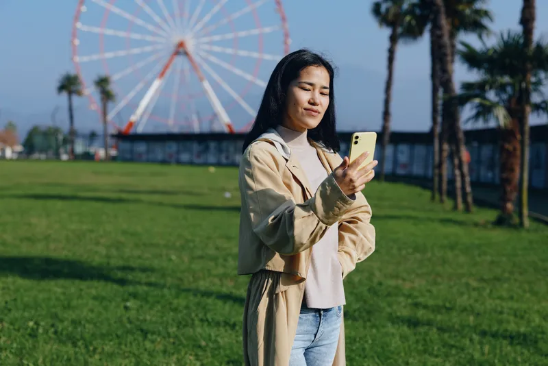 Woman in casual outfit using a smartphone in an outdoor park with a ferris wheel in the background, showcasing leisure, modern lifestyle and urban recreation on a sunny day.