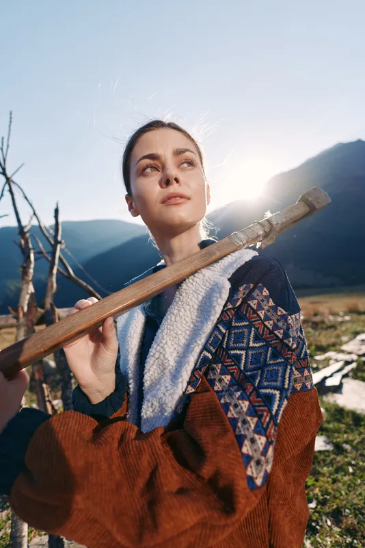 Woman portrait of a confident hiker in patterned jacket against mountains and open outdoors, carrying a wooden stick over shoulder, backlit by warm sunlight for travel lifestyle.