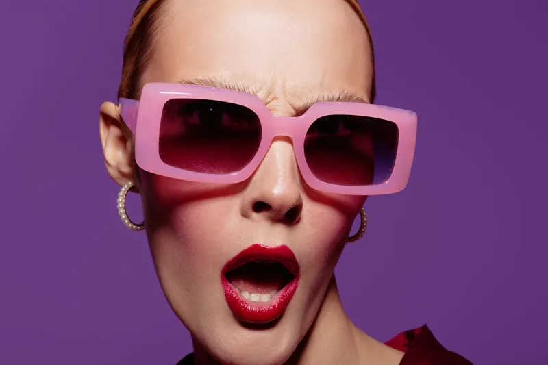 Close-up of a surprised woman wearing large pink sunglasses and red lipstick on purple background. Fashion portrait with expressive facial expression and trendy accessories.