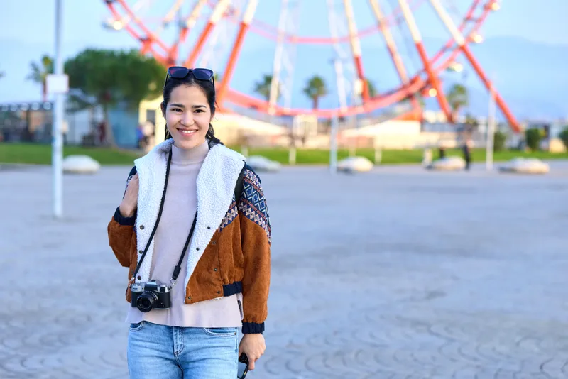 Traveler with camera in a sunny park near a large wheel, wearing a cozy jacket and scarf, capturing casual outdoor leisure moments in an urban setting