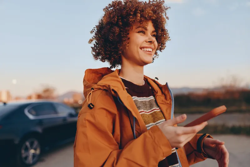 Smiling woman with curly hair in orange jacket stands outdoors near a parked car, looking at a smartphone, relaxed lifestyle moment with warm sunlight, travel vibe during weekend getaway.