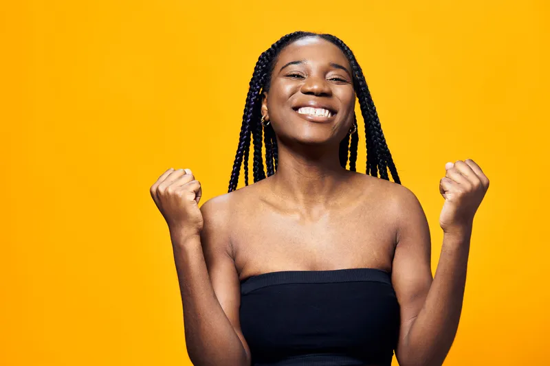 Joyful young woman celebrating success with raised fists against a vibrant yellow background, showcasing emotion and positivity in a stylish black outfit
