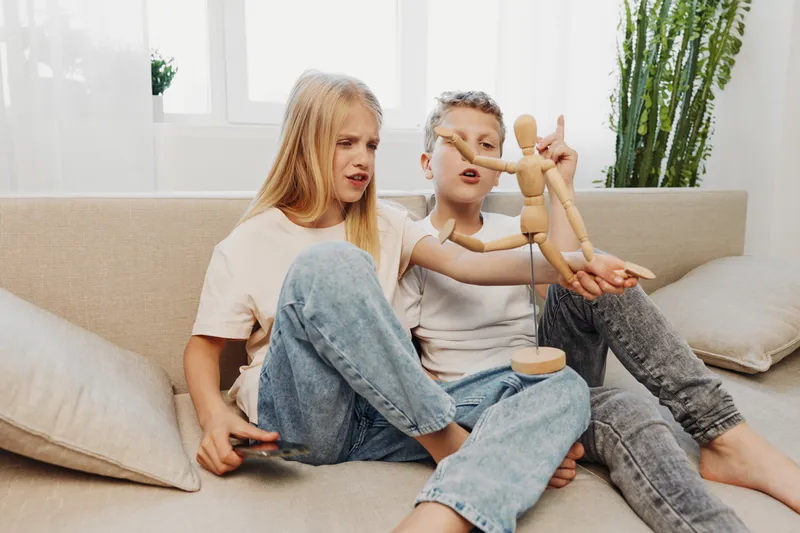 Two children sitting on a couch playing with a wooden mannequin in a bright cozy living room. Concept of childhood play, creativity, and family time.