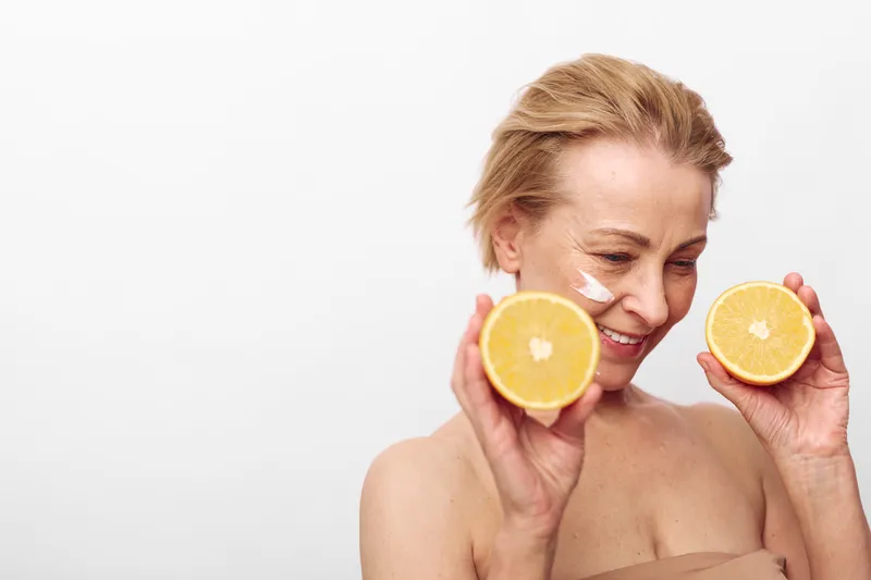 Mature woman with short blonde hair holds two halves of an orange, smiling softly with natural skin and cream on her face, isolated on white background. Concept of skincare and healthy lifestyle.