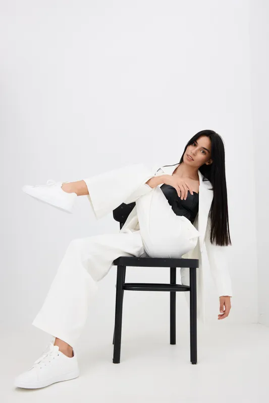 woman sitting on black chair in white pantsuit and sneakers against white background, modern fashion, casual style, relaxed pose