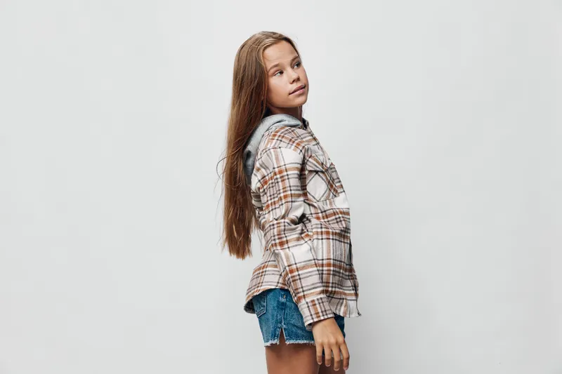 Girl with long hair wearing plaid shirt and denim shorts standing sideways on white background. Child posing with casual outfit for fashion or lifestyle portrait photography.