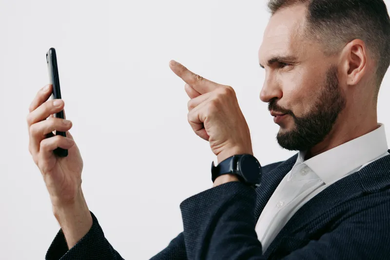 A business professional man sits against an isolated colored background, focusing on a smartphone. He shows a determined emotion, confident posture, and thoughtful expression, conveying leadership,