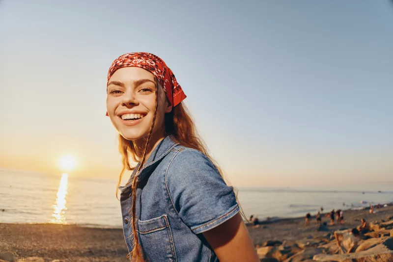 smiling woman in modern boho-western denim street style with a red bandana on the beach at sunset, film color aesthetic, casual and carefree lifestyle vibe