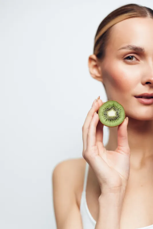Portrait of a woman holding a kiwi near her face, highlighting fresh skincare vibes, natural beauty, and soft bright lighting on a clean white background