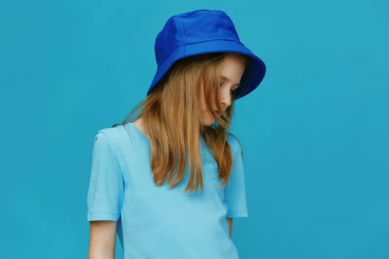 A young female model with long hair wearing a blue T shirt and a blue bucket hat, posing against a vibrant turquoise background, expressing a calm and contemplative mood