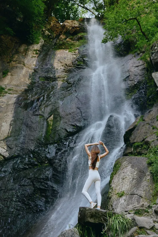 woman standing near waterfall in nature wearing white activewear, serene outdoor scene with rocky cliffs and lush green trees, peaceful and refreshing atmosphere