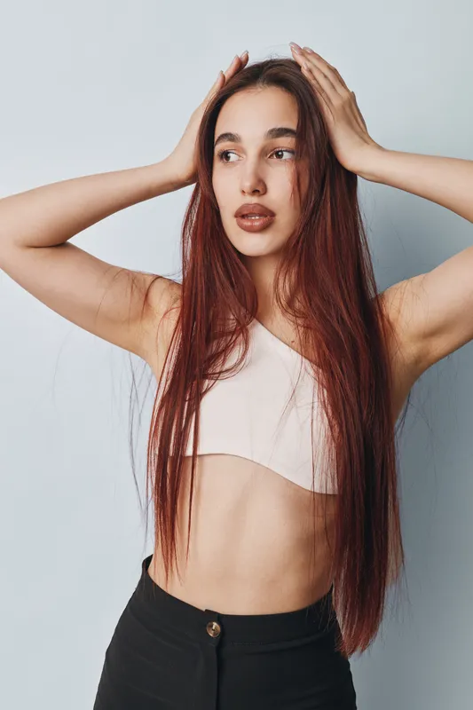 Young woman with long red hair posing with hands on head against white background. Casual outfit with crop top and black pants, natural makeup and thoughtful expression in studio.