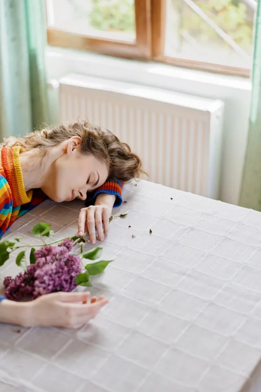 Woman resting at a table with flowers in a cozy home setting, natural daylight and soft textures creating a peaceful, intimate moment of relaxation