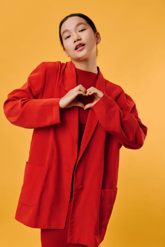 Young woman wearing red coat and sweater making heart shape gesture with hands on yellow background. Asian female expressing love and positivity with casual fashion and confident pose.