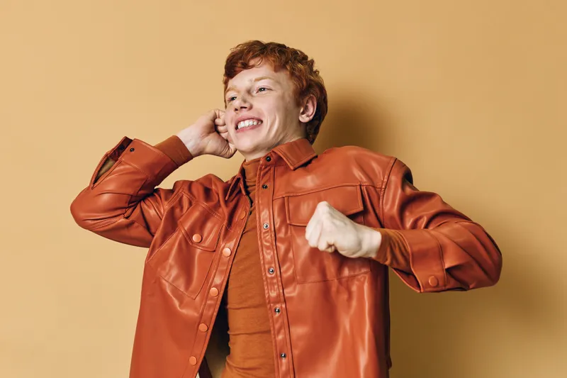 Young man with red hair wearing brown leather jacket smiling happily while posing with fists raised against beige background for positive emotion and style portrait.