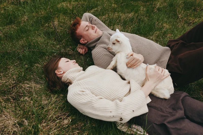 A relaxed couple lounges on the grass in knit sweaters, sharing a tender moment with a curious goat kid nearby. A warm lifestyle scene highlighting affection, companionship, and nature.