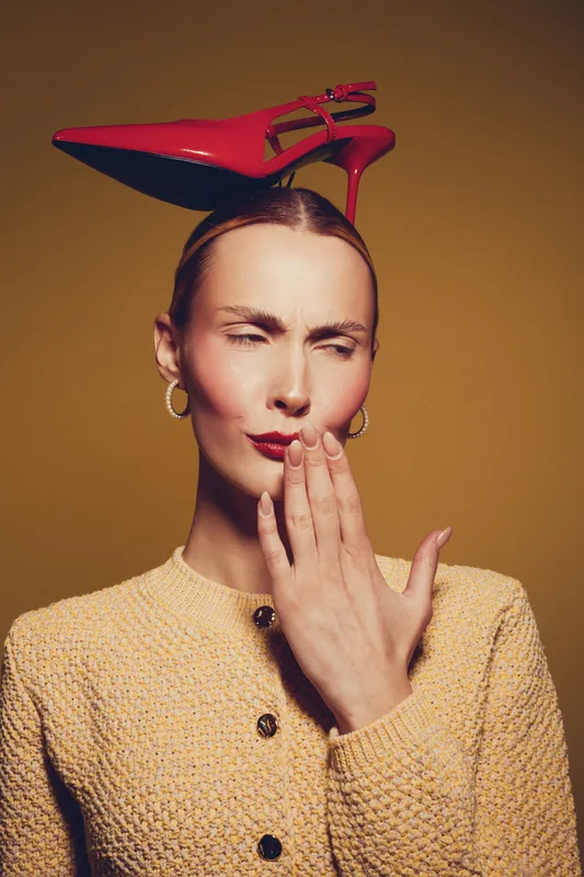 Woman with slicked hair wears yellow textured jacket and balances red high heel shoe on head. She covers mouth with hand and squints eyes against brown studio background.