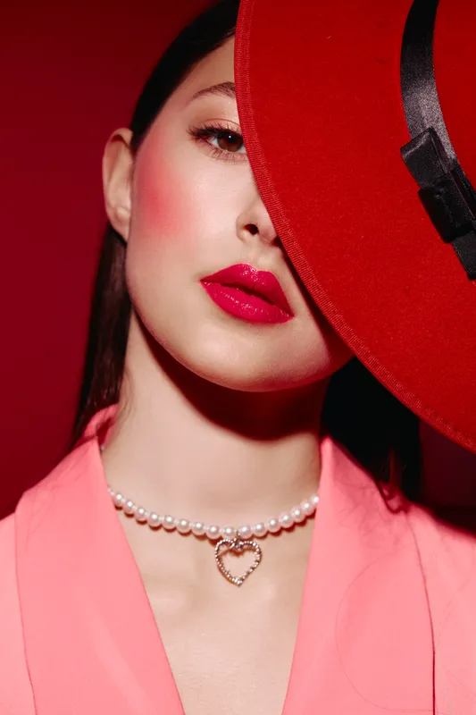 Young woman with striking red lipstick and blush poses stylishly hiding one eye behind large red hat. Bright pink blazer and pearl necklace add elegance. Beauty fashion creative portrait.