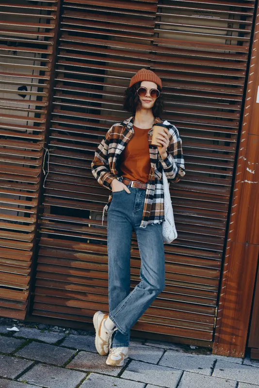 Woman coffee street casual jeans beanie sunglasses plaid leaning against wooden shutter with tote, warm sunlight and urban mood, authenticity, candid lifestyle, golden hour glow and mindful living.