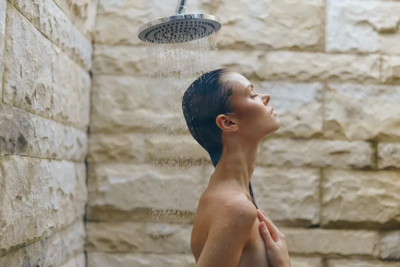 woman showering under modern rain showerhead with stone wall background, calm expression, natural light, wellness and relaxation concept.