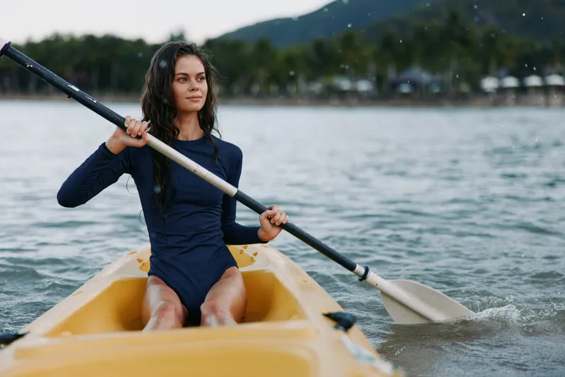 Young woman kayaking in calm water surrounded by forest, wearing navy blue long sleeve swimsuit and enjoying outdoor adventure activity.
