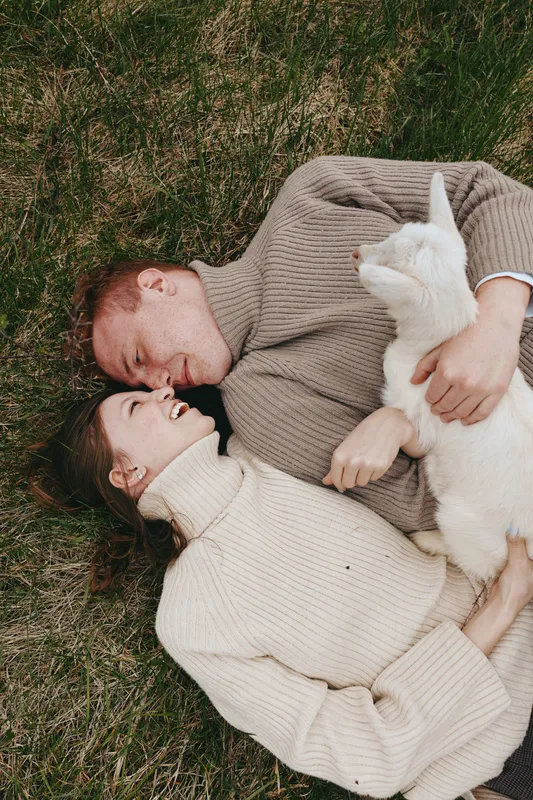 Casual couple lying on grass with a goat kid, enjoying an affectionate outdoors lifestyle moment, showing companionship and cozy bond, natural light adds warmth and calm to the scene
