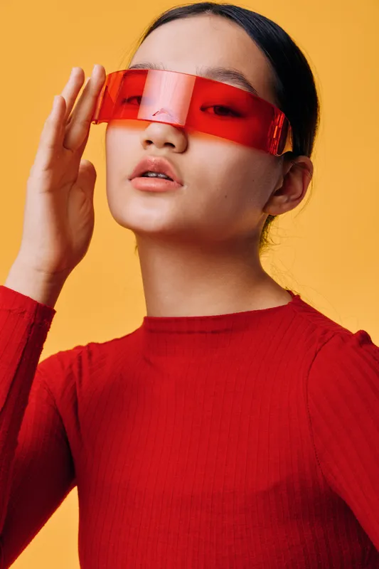 Closeup portrait of woman wearing futuristic red transparent glasses and red sweater, posing against yellow background with confident expression and sleek hairstyle.