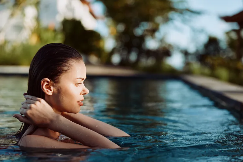 Calm woman in a pool rests her arms on the edge, sunlit water glow, outdoor leisure mood, wellness and self care concept, relaxed atmosphere, natural light, fresh summer vibe