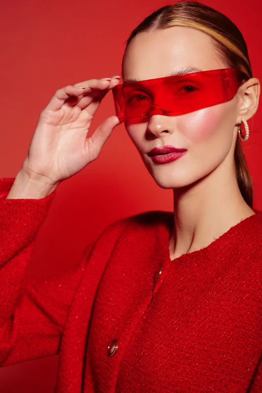 Stylish woman in red sweater and futuristic red sunglasses poses against red background. Fashion portrait with glossy makeup and sleek hairstyle. Concept of modern style and confidence.