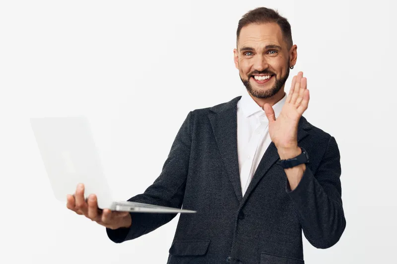 A professional man stands against an isolated colorful background, presenting a laptop while waving with a friendly smile, conveying confidence, approachability, and strong business presence.