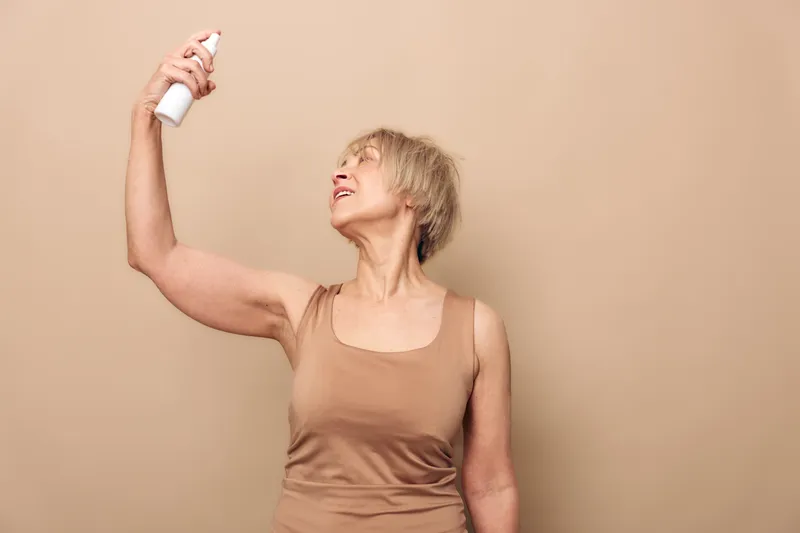 Middle aged woman in beige tank top sprays face mist on skin against neutral background. Concept of skincare routine, hydration, self-care, and healthy lifestyle for mature skin.