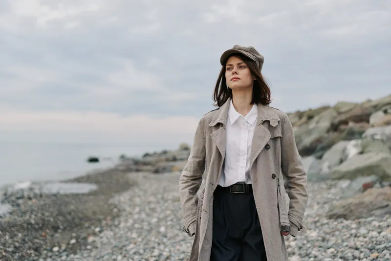Young woman in vintage coat and cap walks on rocky beach under cloudy sky. Casual autumn fashion style with natural makeup and confident expression by the sea shore landscape.
