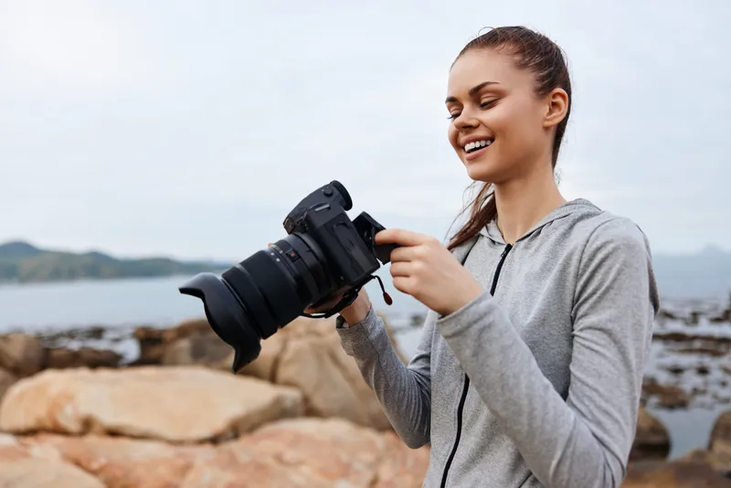 Young female photographer enjoying nature, holding a camera on rocky beach, smiling, showcasing passion for photography in a serene coastal setting