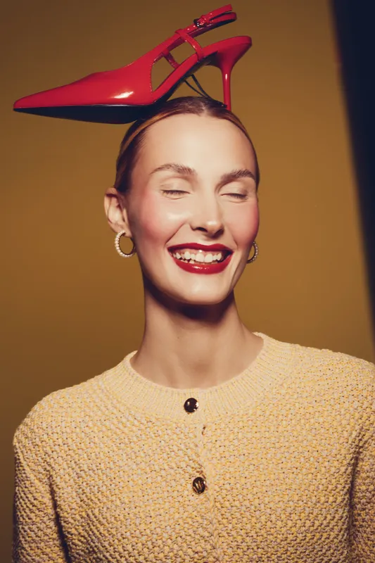 Smiling woman with closed eyes wears red high heel shoe on head, dressed in textured beige sweater with buttons. Concept of fashion, creativity, and playful style on warm background.