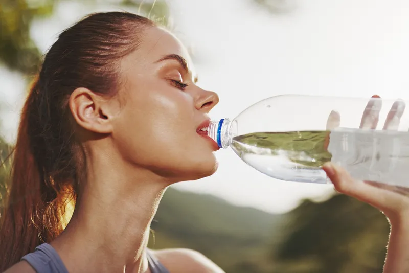Young woman drinking water from a plastic bottle, enjoying refreshing hydration in a lush outdoor setting, representing healthy lifestyle and comfort