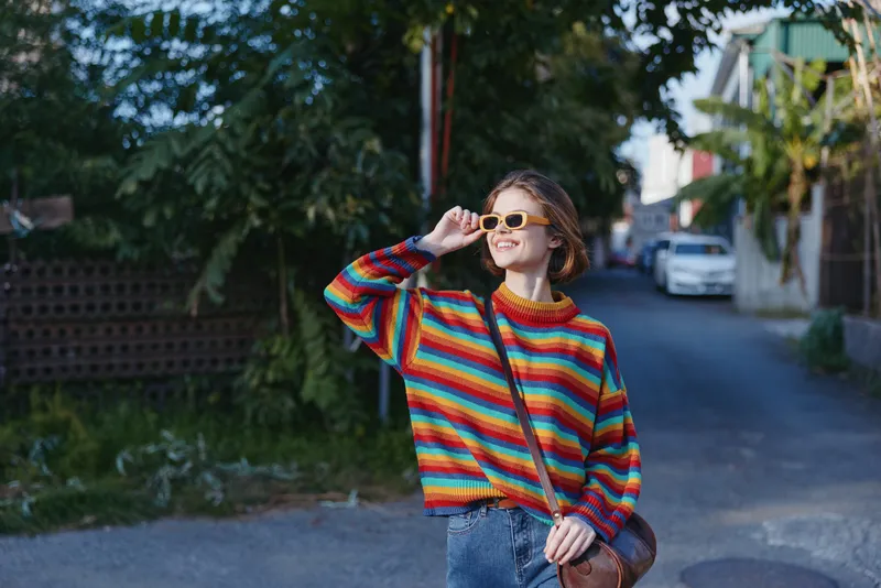 Young woman in her 20s wearing a striped sweater and sunglasses on a sunlit street, smiling in casual outfit with short hair and crossbody bag, walking in a tropical neighborhood.