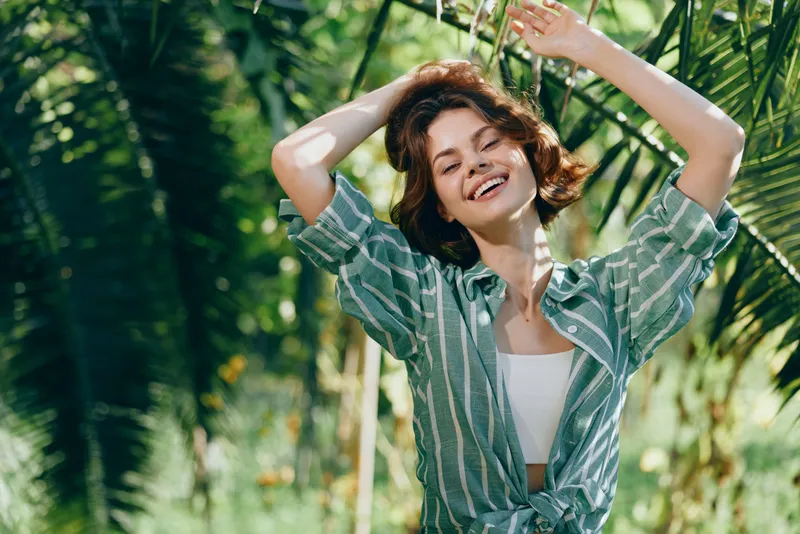 Joyful woman in a striped shirt raises arms amid tropical greenery and sunlit smiles.